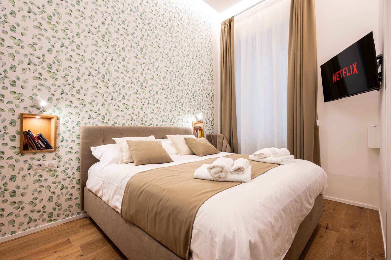 Modern bedroom with leaf-patterned wallpaper, beige and white bedding, and a wall-mounted Netflix TV.