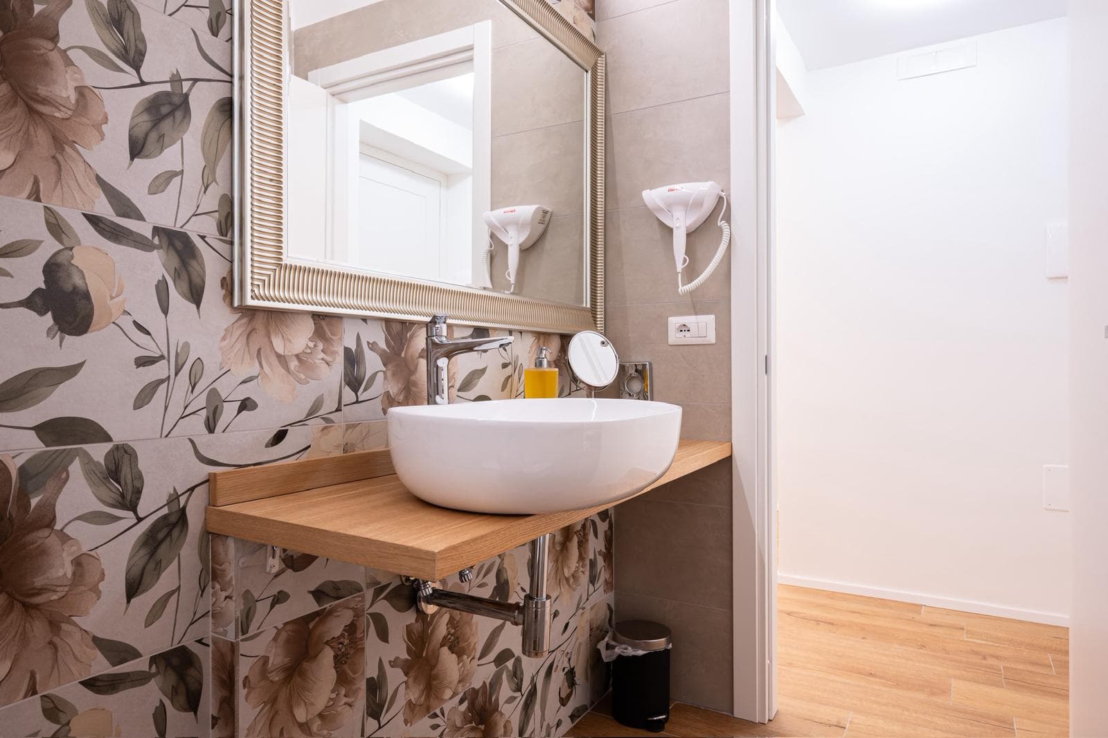 White vessel sink on a wooden countertop against floral wallpaper in a modern bathroom.