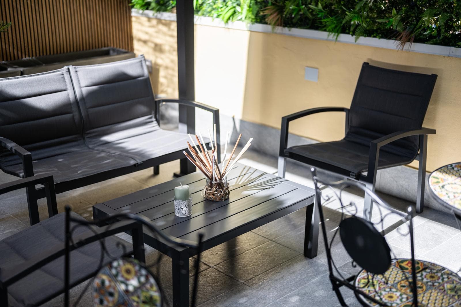 Sunlit outdoor patio featuring black metal furniture, a coffee table, candle, and reed diffuser.