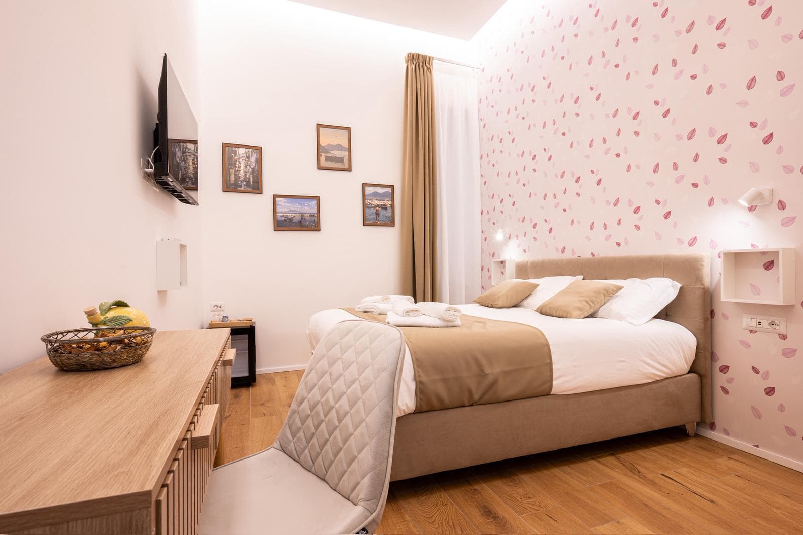 Modern bedroom featuring pink leaf wallpaper, a beige upholstered bed, wooden desk, and grey chair.